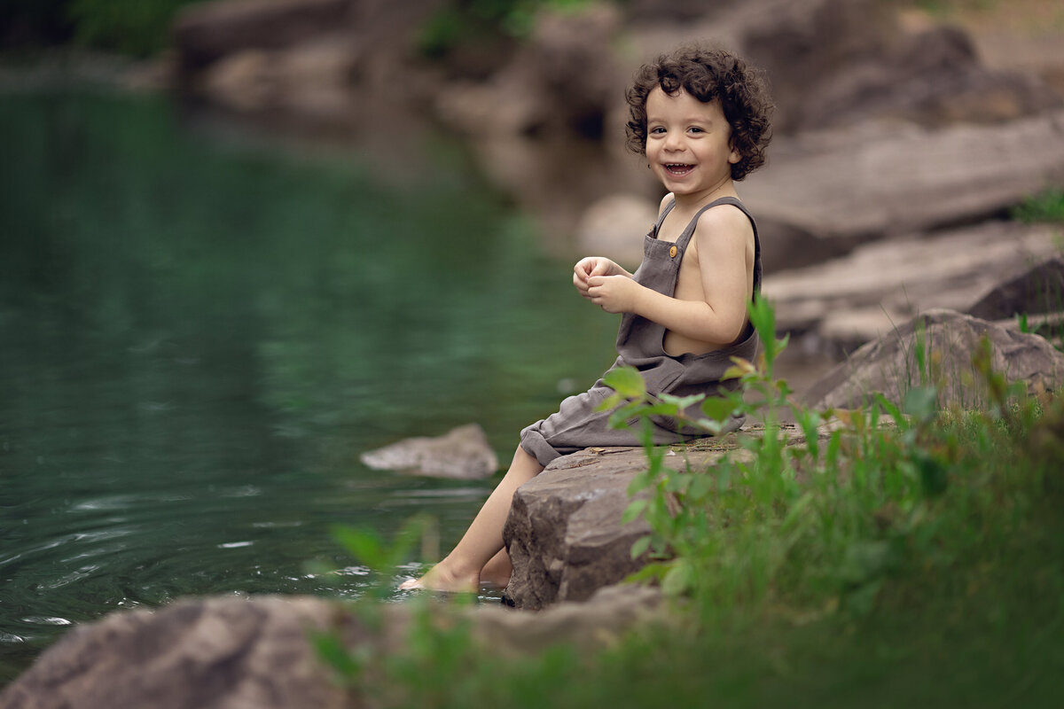 A young toddler boy in brown overalls sits on a rock with his feet in the water smiling for a New Jersey Family Photographer