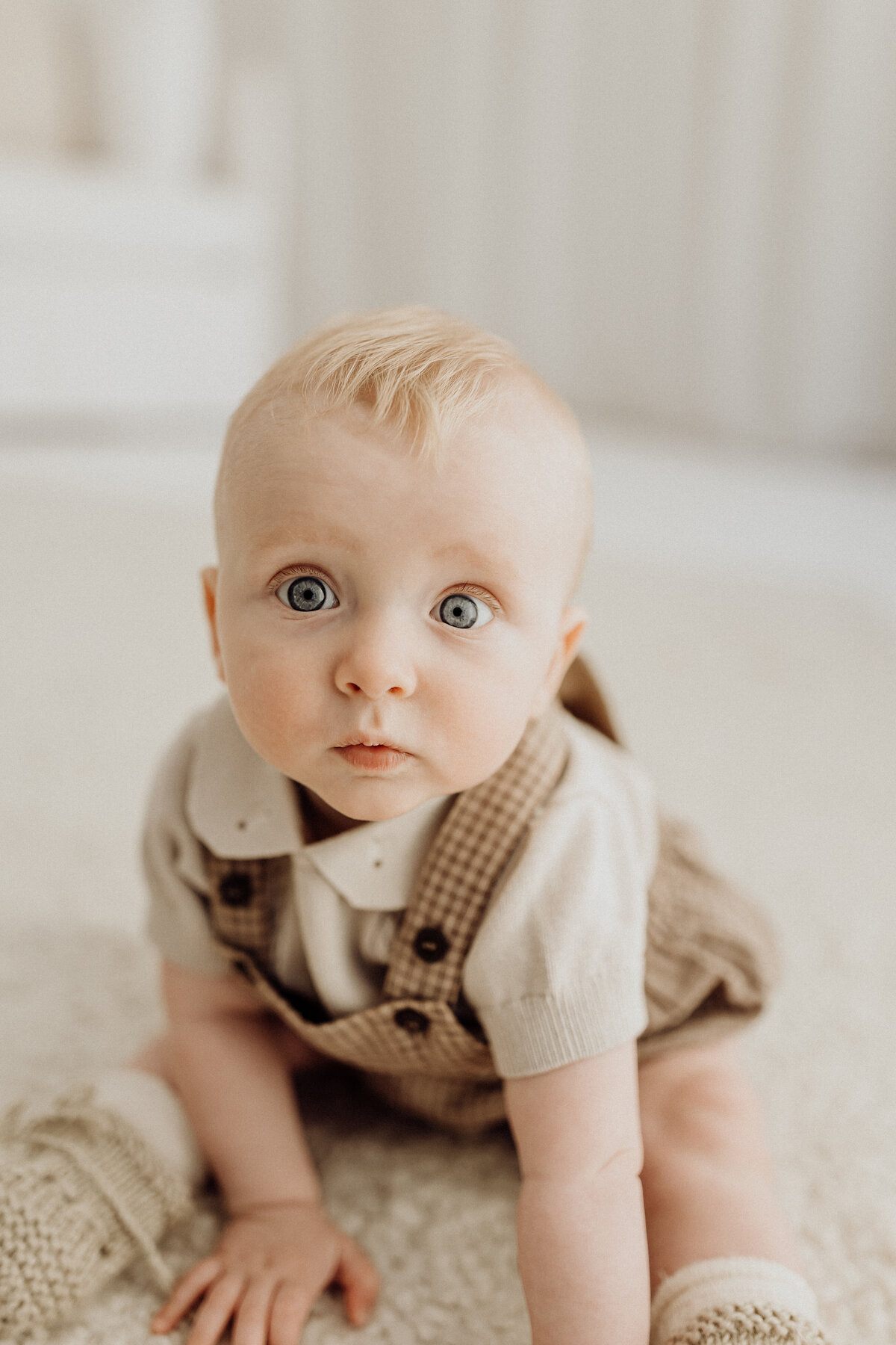 A little baby boy wearing a cute checked overall and knit top and looking at the camera during his milestone photoshoot in lucy janes photography studio in christchurch