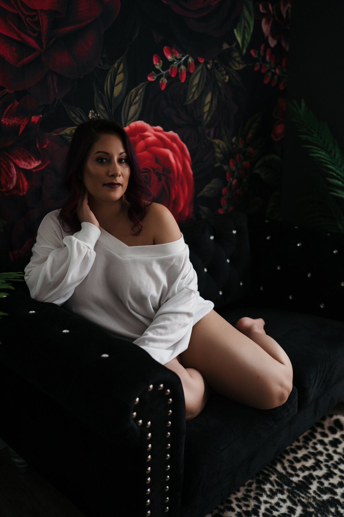 A woman in a white oversized shirt sits across a black couch in a studio in front of a floral tapestry