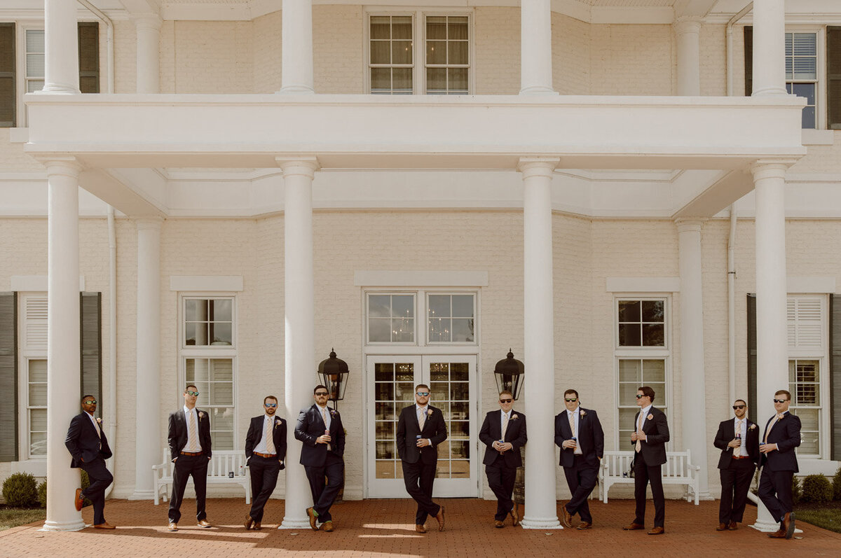 Groomsmen stand in a beer in hand in front of wedding venue main doors
