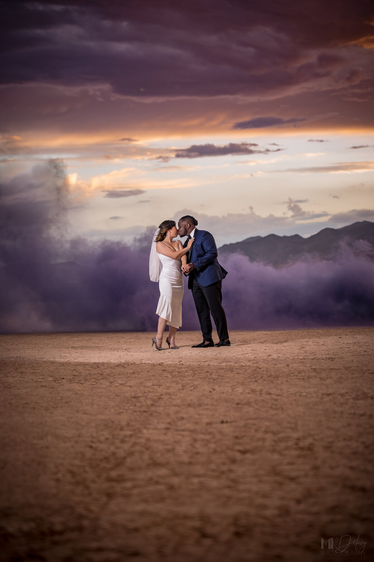 Bride kisses her groom in the desert with purple smoke bomb in the wind las vegas elopement on the dry lake bed  at golden hour groom in blue suit jacket and black  pants  las vegas elopement eloping in vegas  las vegas wedding photographers las vegas wedding photography mk delacy photography