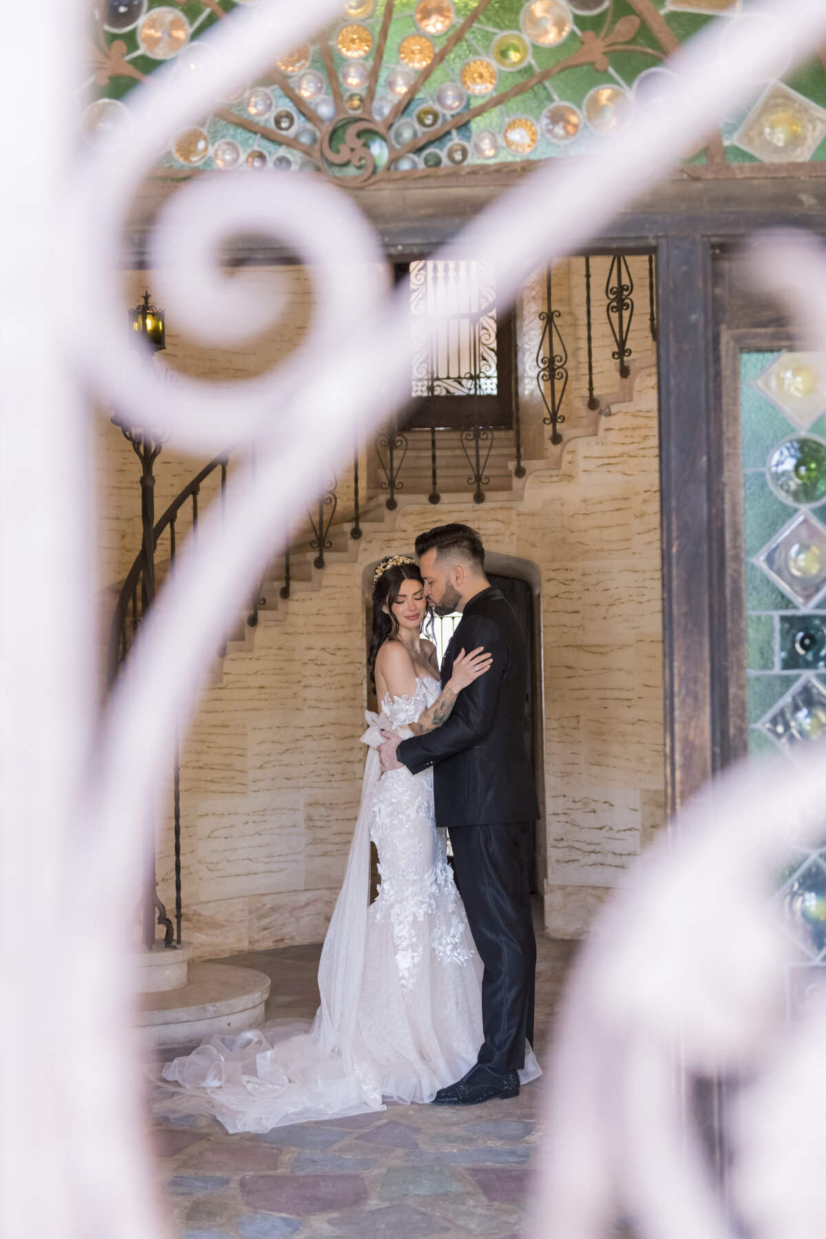 A bride and groom share a romantic embrace inside a stunning architectural venue, framed by intricate ironwork. Wedding coordinated by WINK! Weddings.