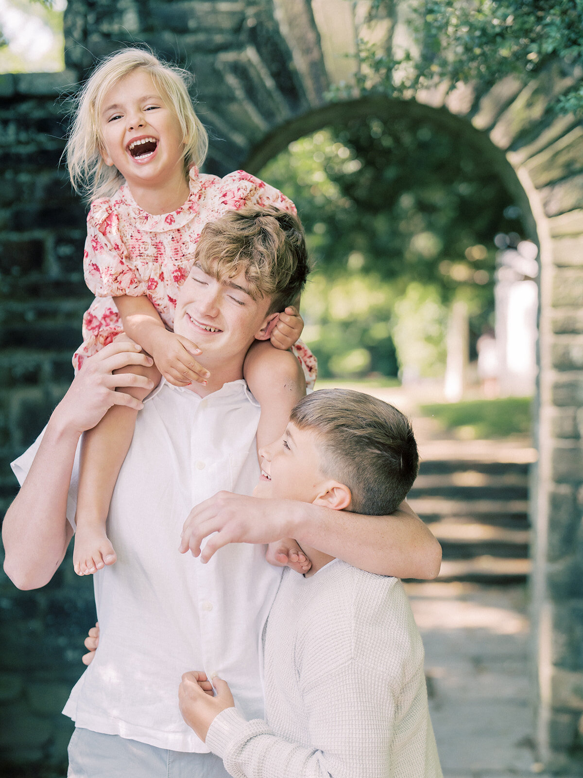 Little girl sits on her big brother's shoulders and laughs as a younger brothers hugs the bigger brother, too.
