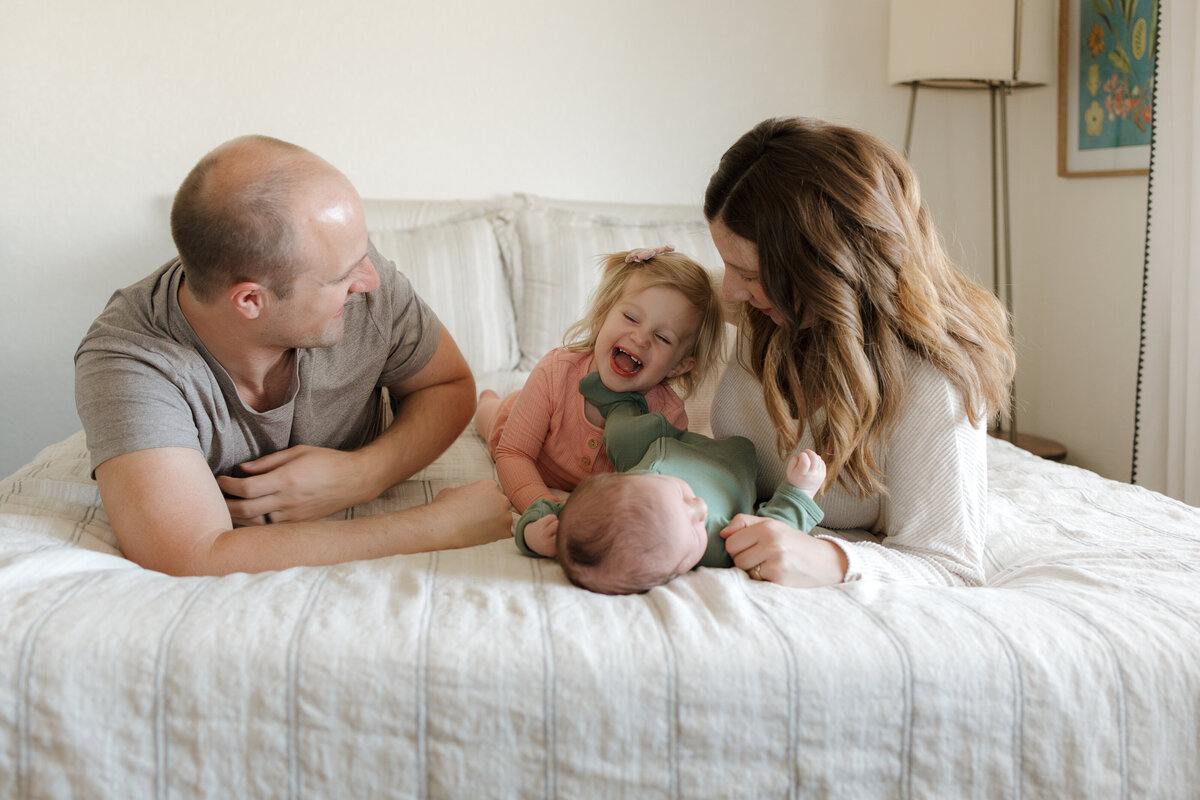 Longmont family laying on stomachs on bed. Toddler is laughing while newborn is putting his foot on her neck accidentally