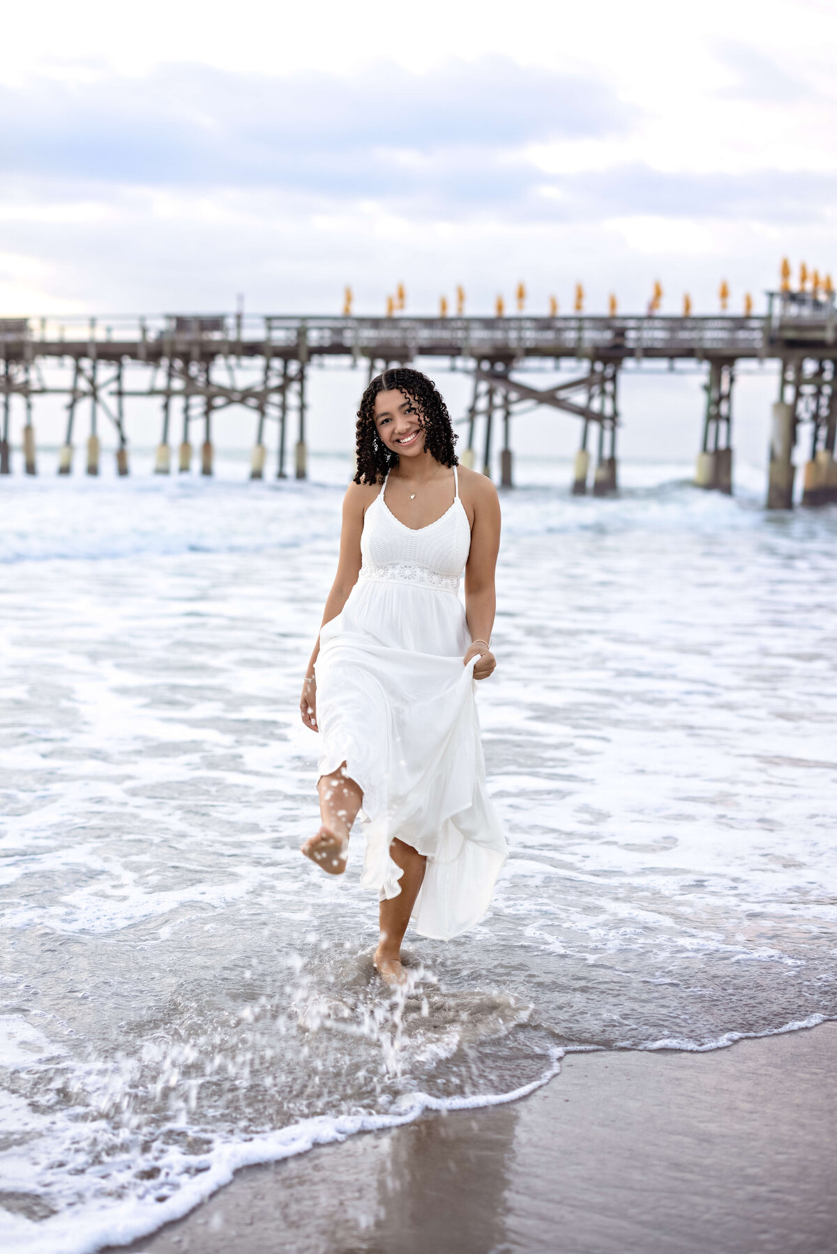 Senior girl kicking up ocean water near the Cocoa Beach Pier, smiling with playful energy and a wind-blown dress.