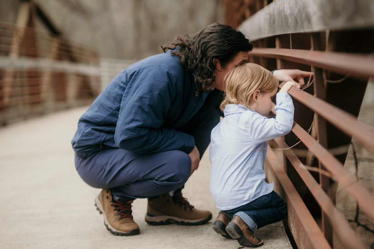 Father and son kneel on a bridge looking through the railing.
