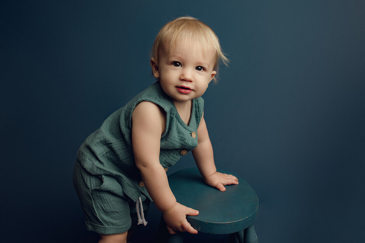 a one year old boy holding a blue stool on a blue backdrop