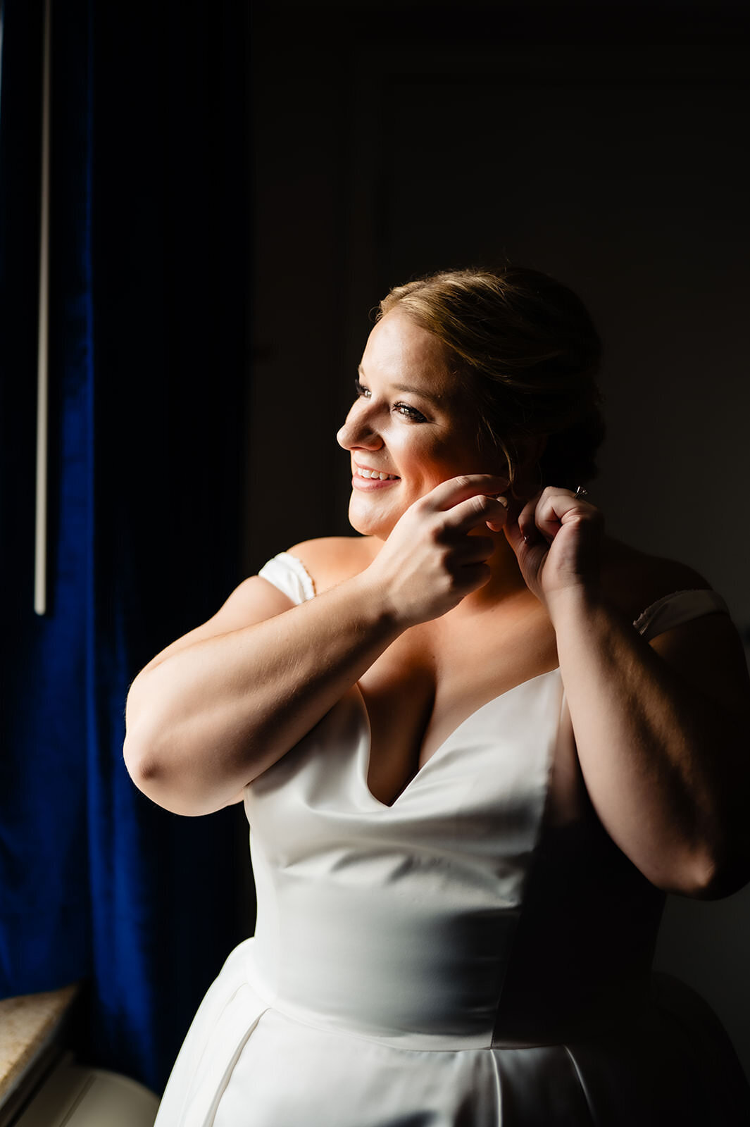 A radiant bride in a white dress, smiling as she puts on her earring, with light streaming in from a window