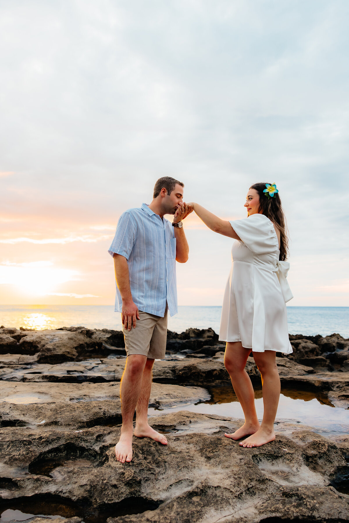 Couple sharing a sweet moment on the rocks at sunset in Oʻahu, with him kissing her hand at Paradise Beach Cove celebrating their wedding anniversary