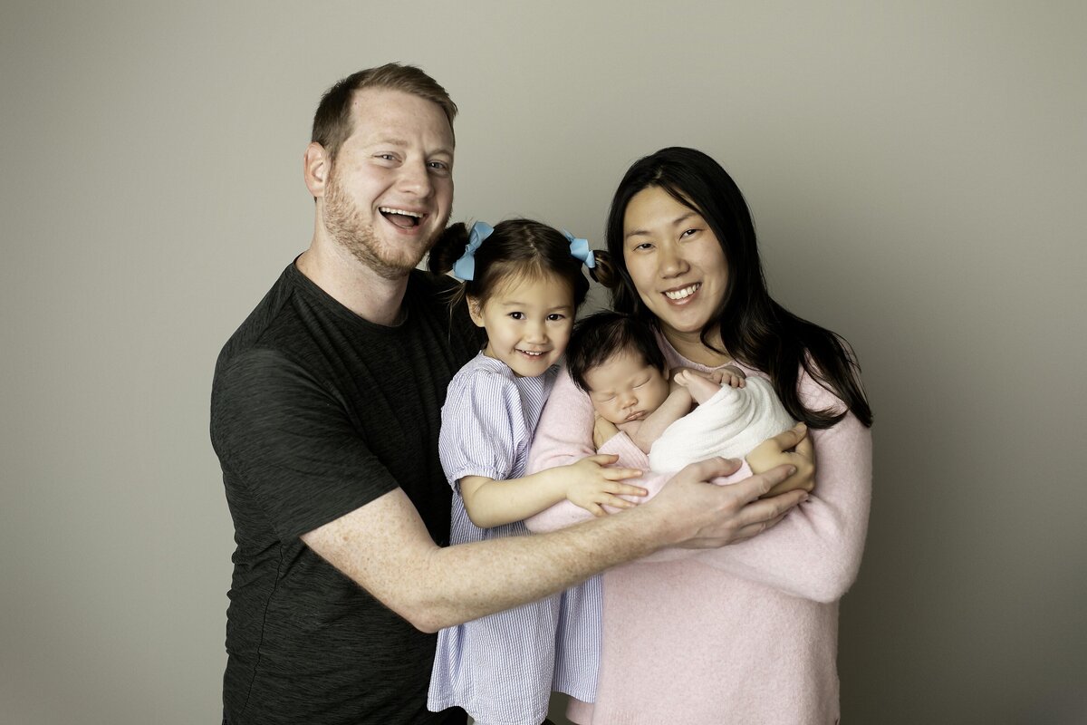 A loving family of four posing together, with mom gently cradling the newborn baby while dad and the older sibling embrace them, capturing a warm and heartfelt family moment
