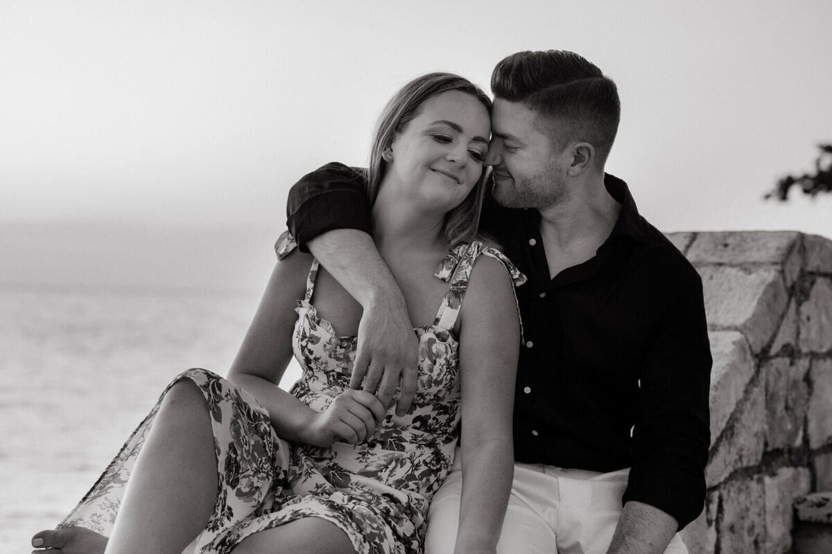 Black and white photo of the engaged couple, touching heads, sitting on a stone, fronting the ocean, in Round Hill Hotel, Jamaica.