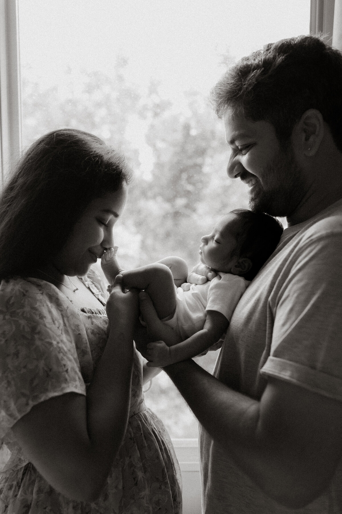 Black and white photo of new parents holding their baby together by the window during an in-home newborn session.