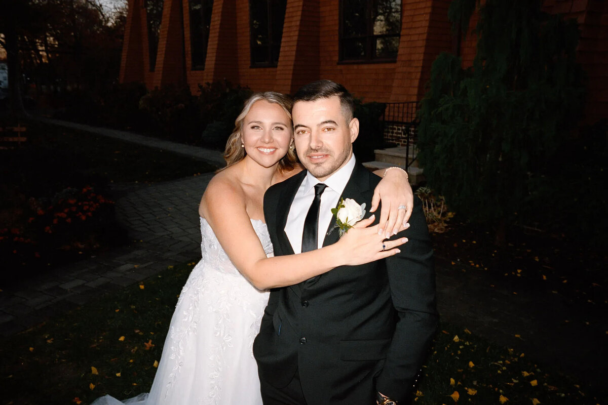 A bride in a white dress hugs a groom in a black suit and tie outdoors, both smiling at the camera. Captured by an experienced NJ wedding photographer, they stand on a path beside a building with brown shingles and greenery.