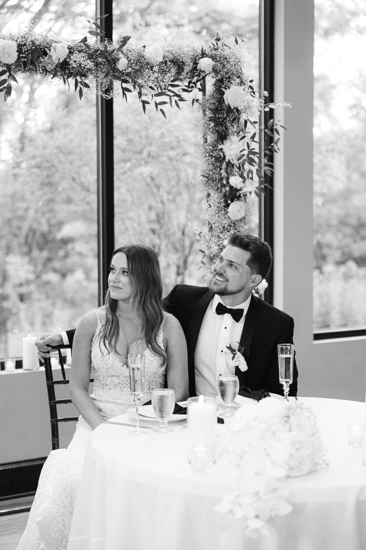 Bride and groom share candid moment at reception table under floral arch in black and white.