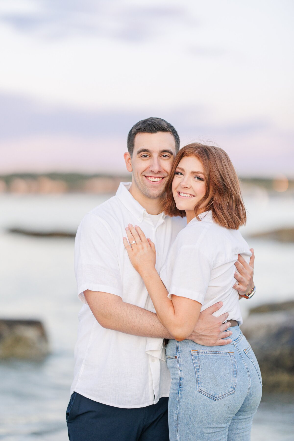 An engaged couple hugging and smiling for the camera at sunset at Wingaersheek Beach.