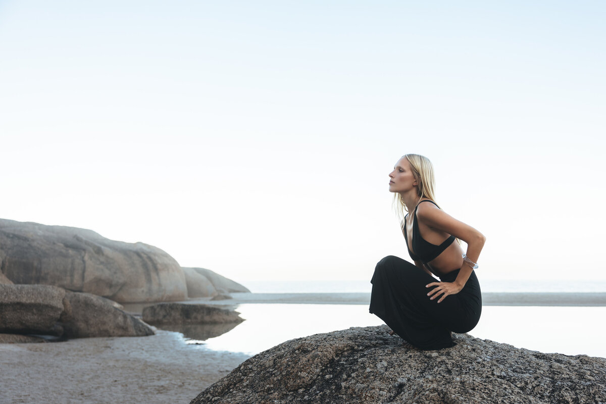 Model Johanna hurkt krachtig op een rots in Kaapstad, gekleed in een zwarte cut-out jurk, met de blik gefocust richting horizon en omringd door ruige rotspartijen en rustig kustwater.