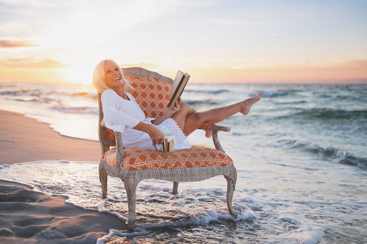 Older woman with white hair wearing a white long-sleeve dress sits on an antique chair on a Florida beach at sunset while reading a book.