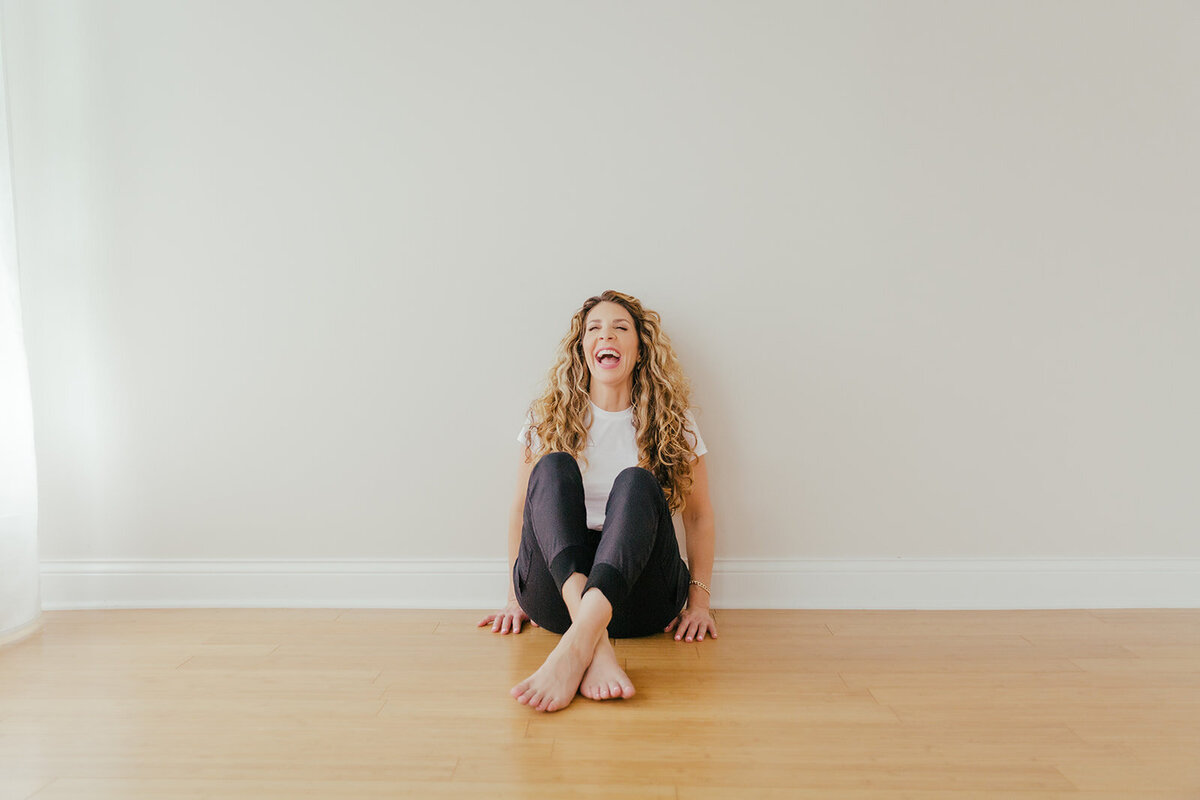 woman in white tee and black sweats in sitting on a hardwood floor and leaning against a white wall and laughing.