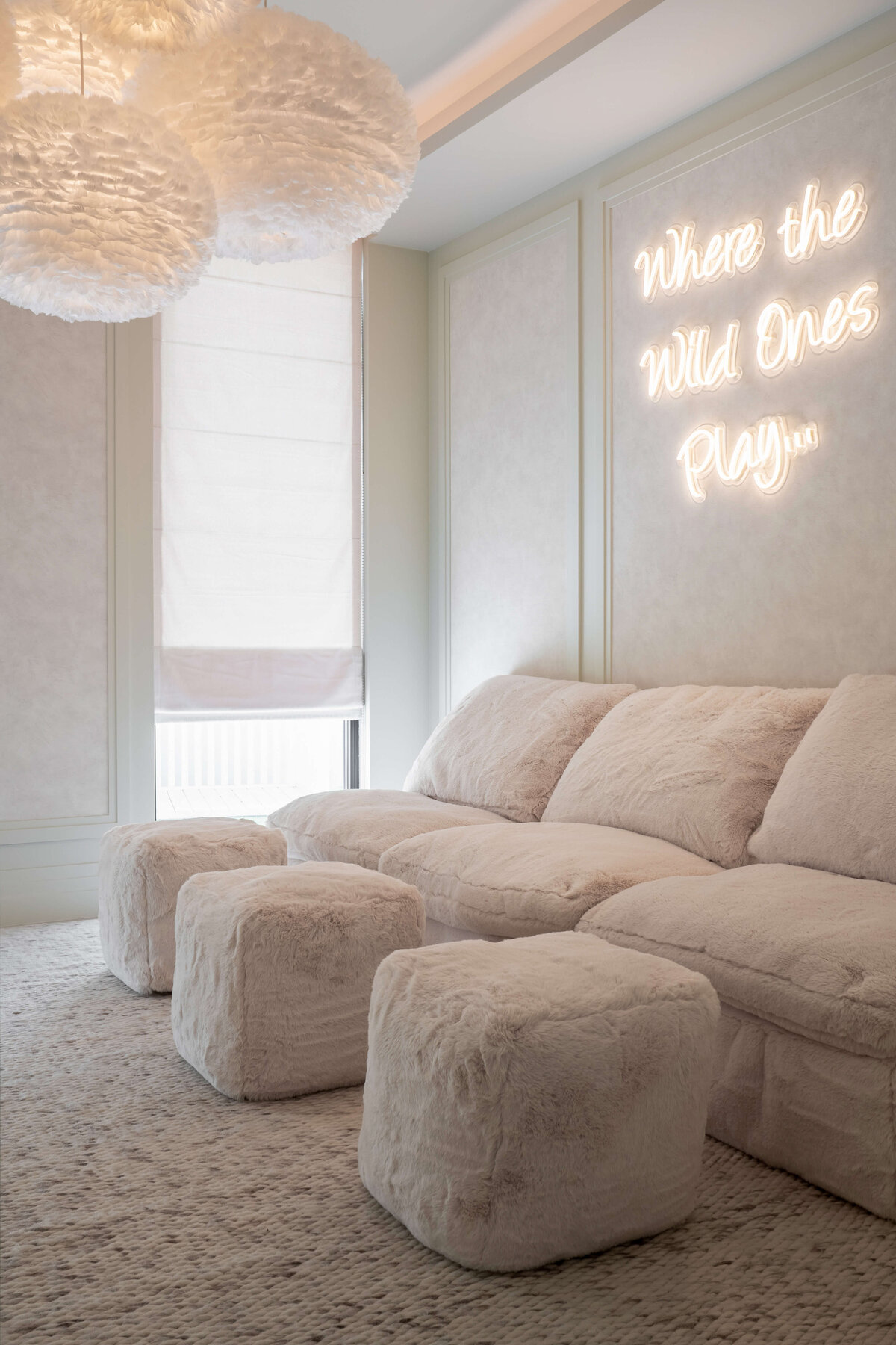 Spacious playroom with a light, airy aesthetic. There is a large, plush tan couch with three fuzzy pouf ottomans. Behind the couch is a softly illuminated neon  sign reading 'Where the Wild Ones Play...'. Above are four white, feathery spherical pendant chandeliers.