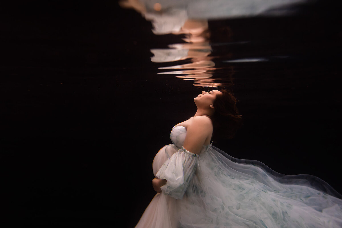 A serene underwater maternity photo featuring a woman in a flowing floral gown, her head tilted back gracefully as her reflection shimmers above. Captured by a Dallas underwater maternity photographer, this ethereal scene emphasizes the gentle elegance of motherhood, with fabric floating weightlessly in the dark water.