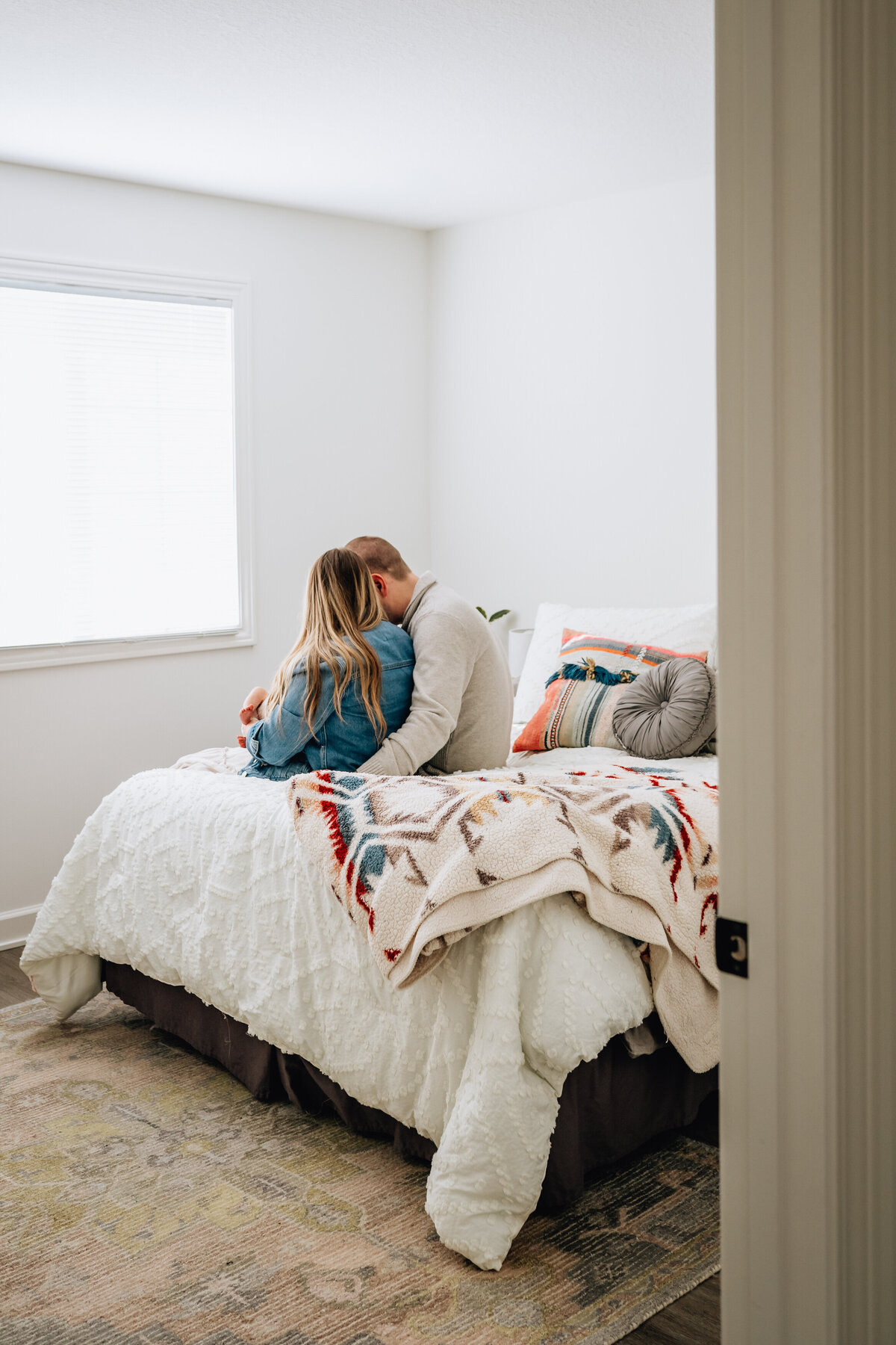 A wide-angle shot of a couple sitting closely together on the edge of a bed in a minimalist bedroom, facing away from the camera. The room is bright, with natural light streaming in from a window on the left