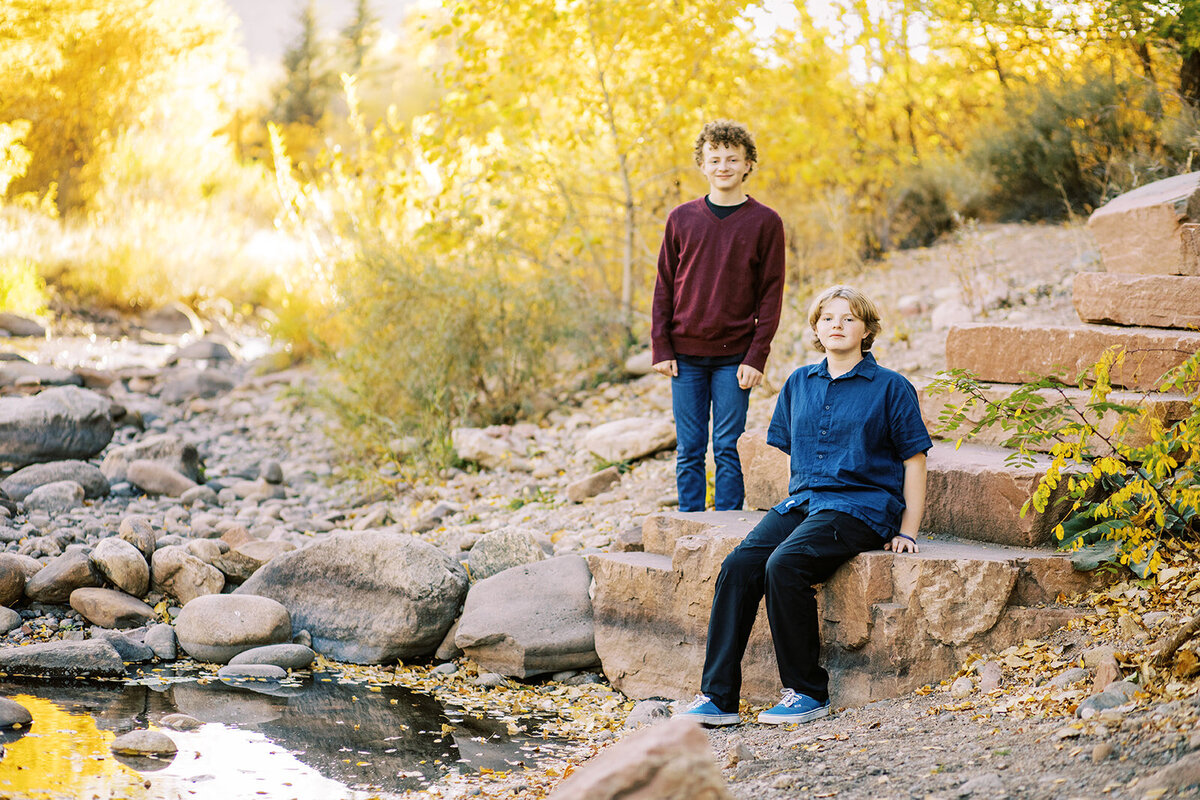 two brothers stand and sit on flagstone rock steps by a river on a fall day in lyons, colorado