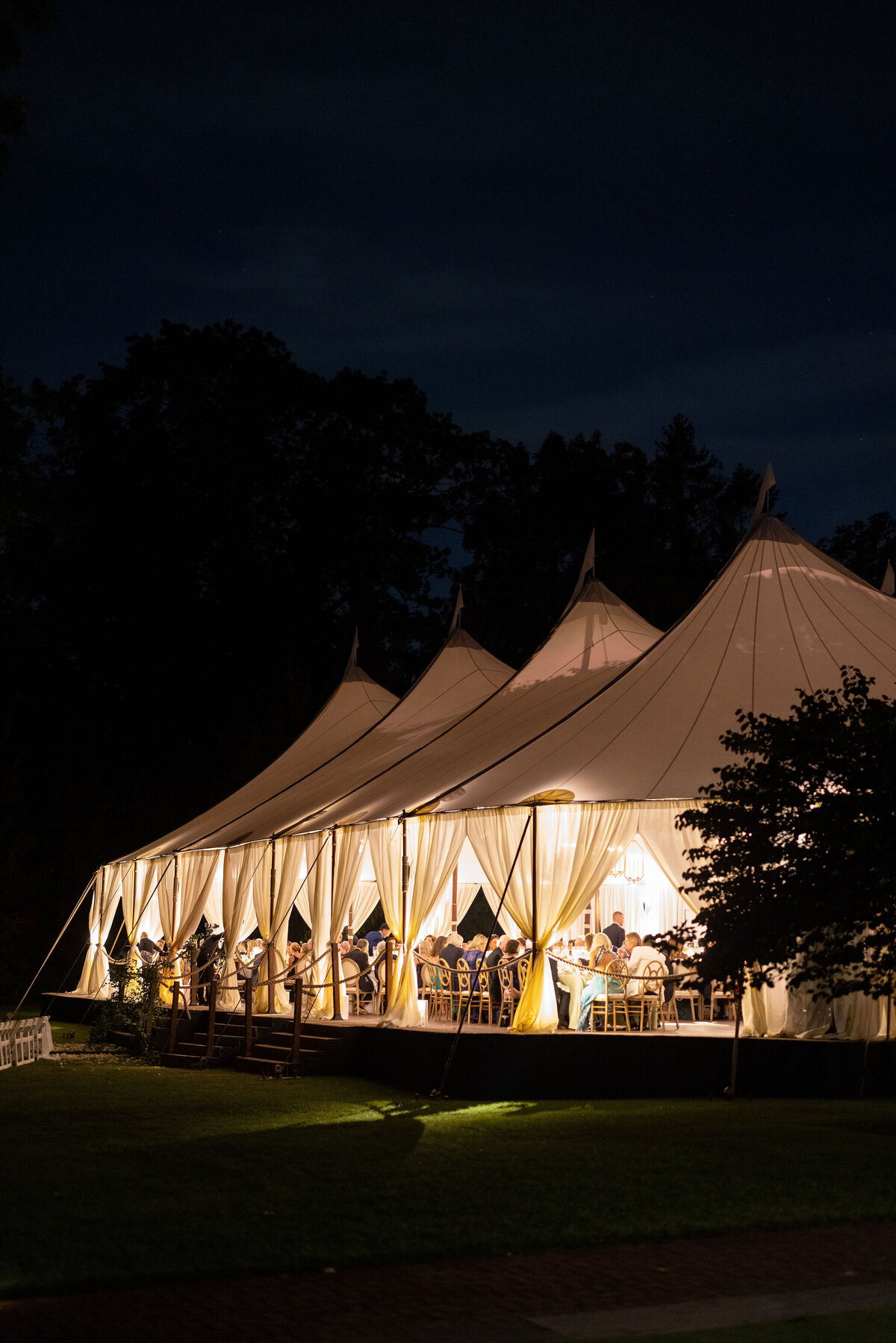 Nighttime view of a large, illuminated wedding tent on a manicured lawn, glowing warmly against the dark evening backdrop.