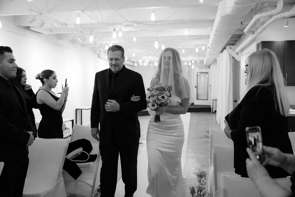 A bride in white and her father walk down a small aisle of her intimate wedding ceremony as loved ones look on. 