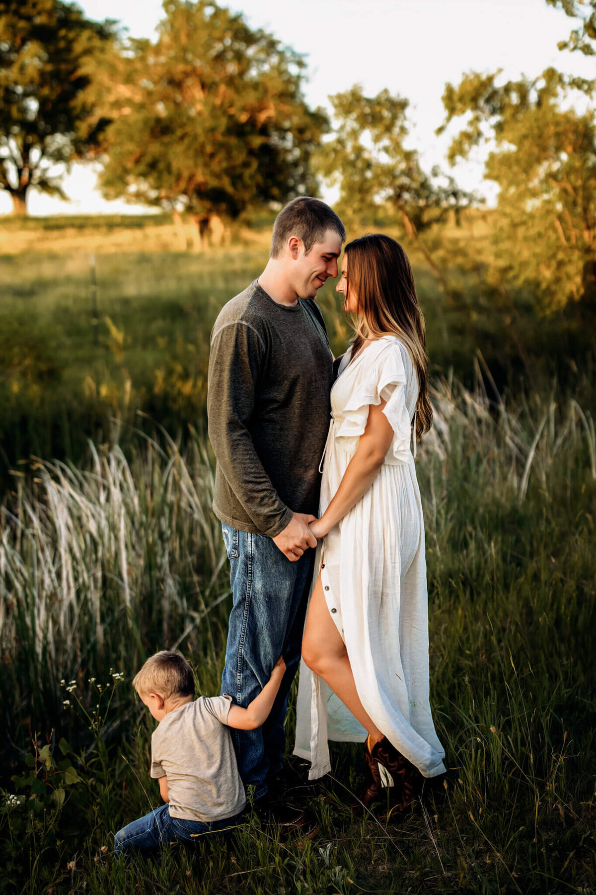 A couple stands close together, holding hands and smiling at each other in a grassy field. A young boy, sitting on the ground, holds onto the man's leg. This sweet outdoor portrait captures a genuine family moment among trees and tall grass.
