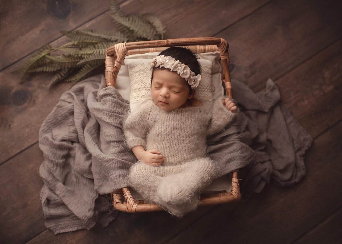Best Newborn Photographer | Aerial image of baby sleeping on rattan bed in styled cream newborn photography outfit with headband. Greenery in background.