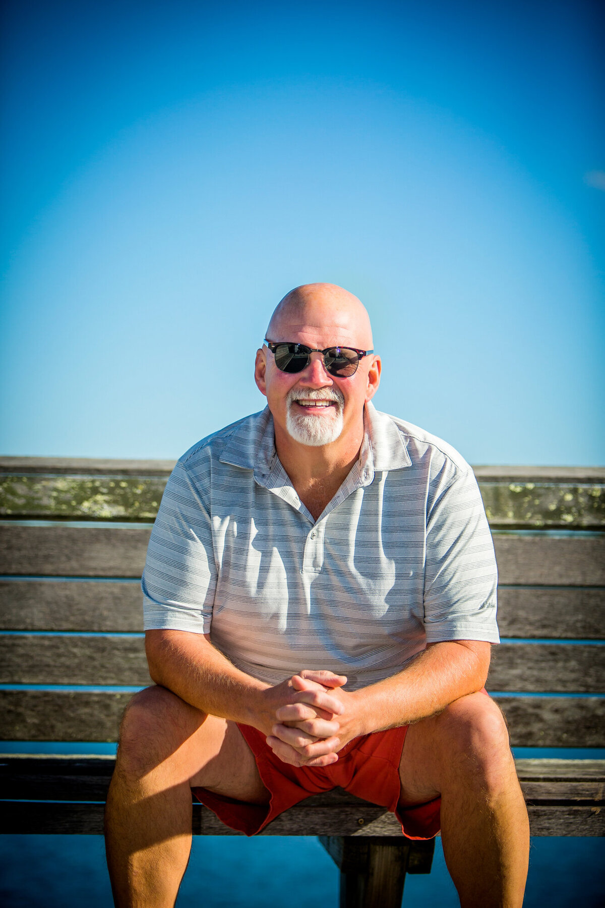 A male dating portrait client sitting on his deck with a short-sleeved shirt, shorts & sunglasses