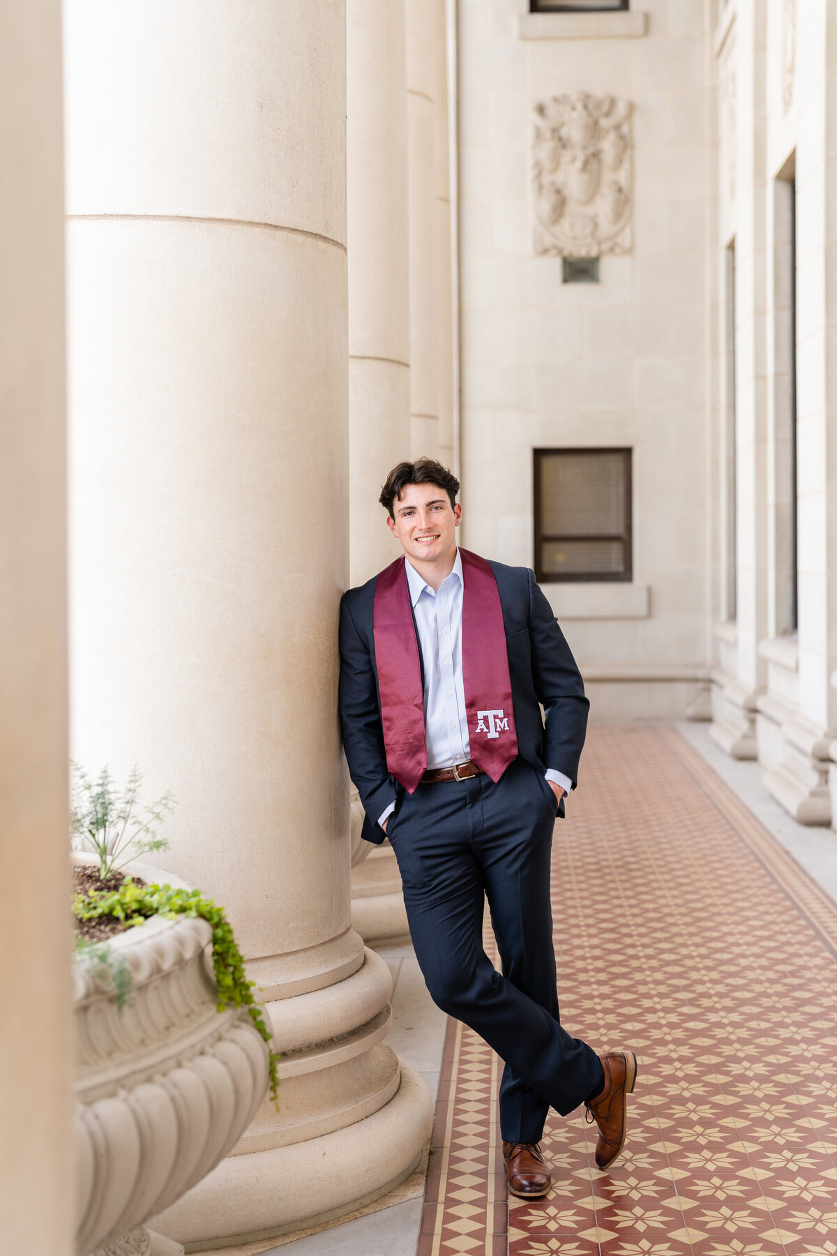Texas A&M senior guy leaning against column with hands in pocket wearing a suit and maroon stole in the columns of the Administration Building