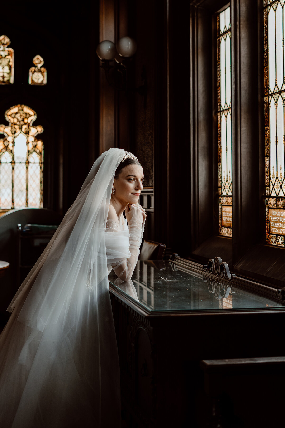 Bride in a classic wedding dress and veil gazes thoughtfully out of a stained glass window in a dimly lit room, capturing a serene and reflective moment