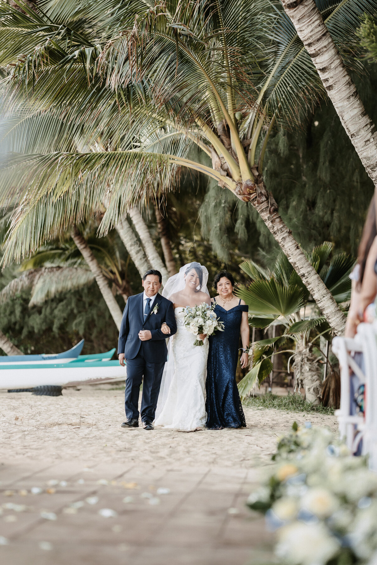 Bride walking down the aisle with parents arm in arm palm tress in the background on the beach