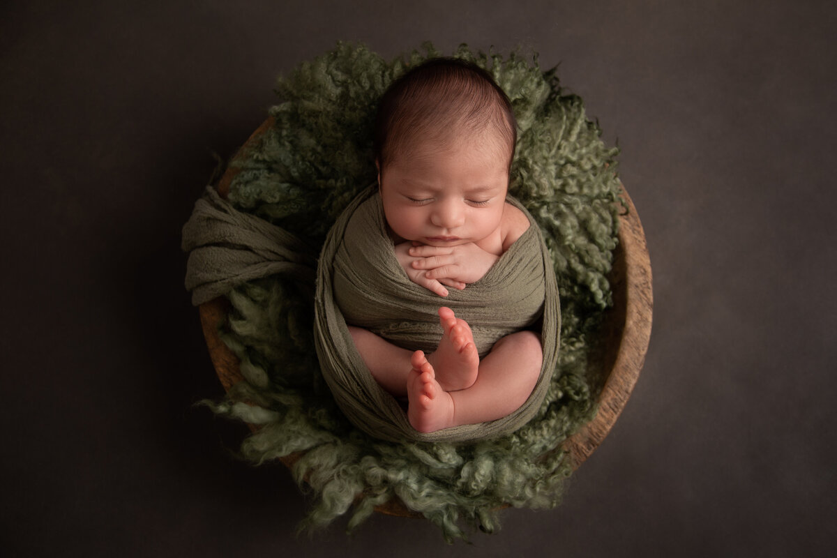 Sleeping baby wrapped in dark green on matching fur in a wooden bowl on a grey background.