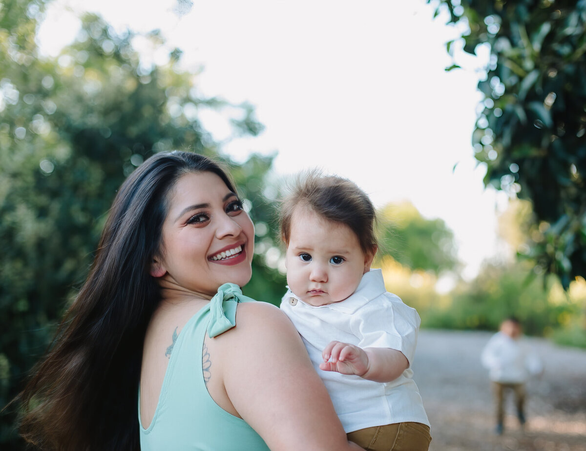 Long Beach FamilMom holding her infant son while her oldest child plays in the back at WillowSpring Park