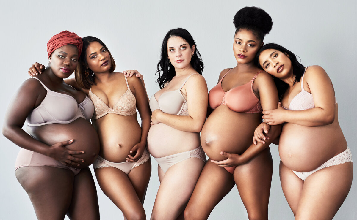 A group of diverse women are dressed in their undergarments, holding their bellies, and posing.