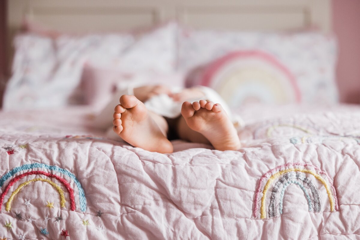 A person's bare feet sticking out from under a quilt with rainbow designs on a bed, captured by a renowned family photographer Pittsburgh.