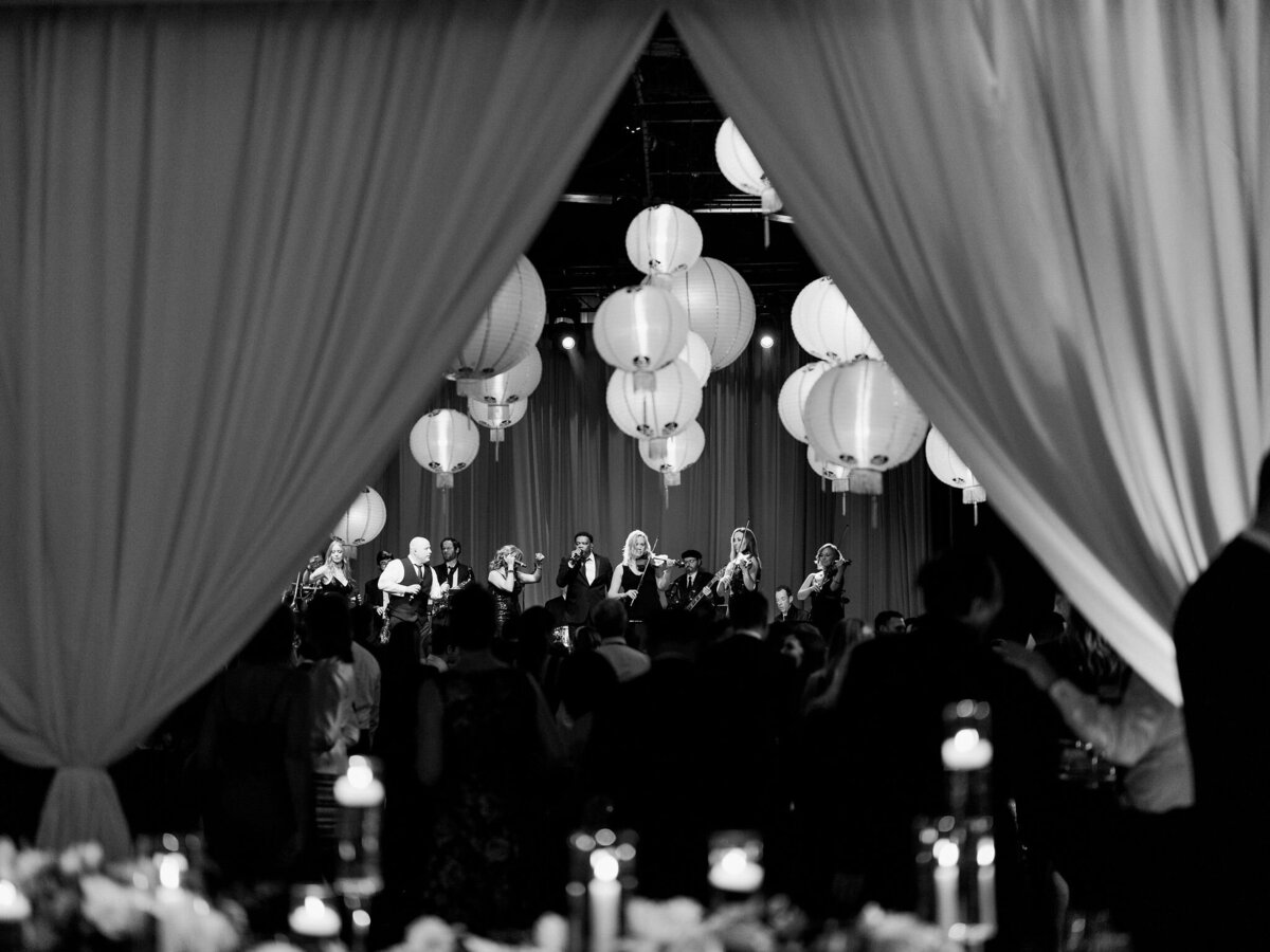 A live band performs on stage, framed by draped curtains and hanging lanterns, creating a lively atmosphere.