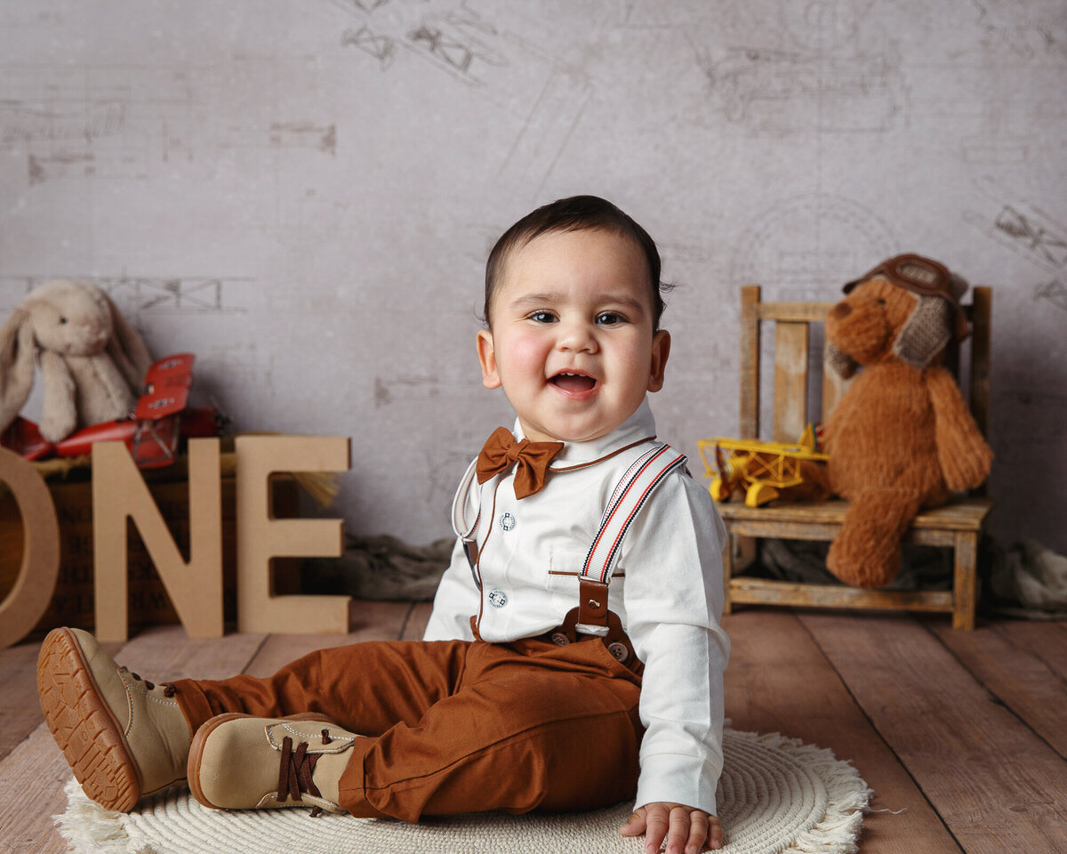 One year old milestone portrait of baby boy wearing brown pants, suspenders and bowtie with teddy bears in the background