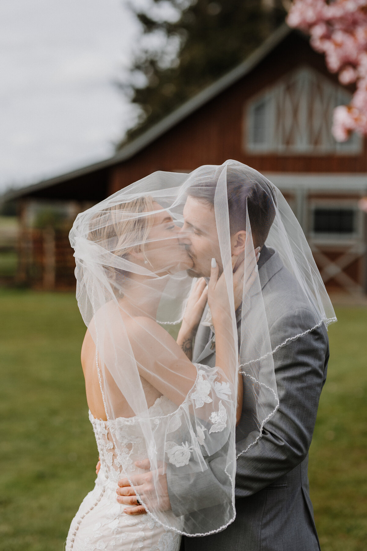 Bride and groom kissing underneath a veil in front of a barn