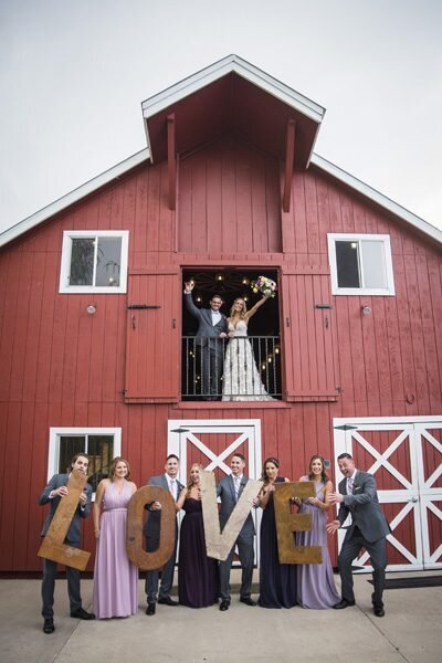 The bride and groom stand in the upstairs doorway of a red barn while their wedding party stands on the ground below holding the letters L-O-V-E.