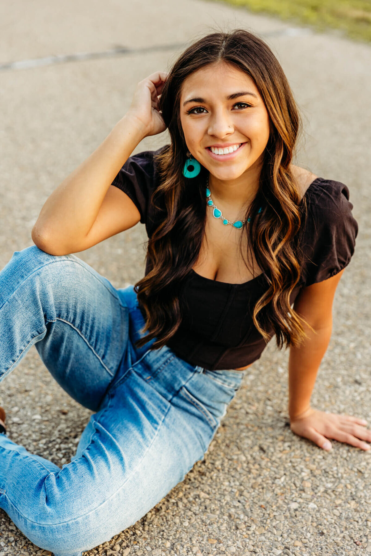 gorgeous young lady in high school sitting on a road and looking up as she rests her elbow on her leg for her senior photos