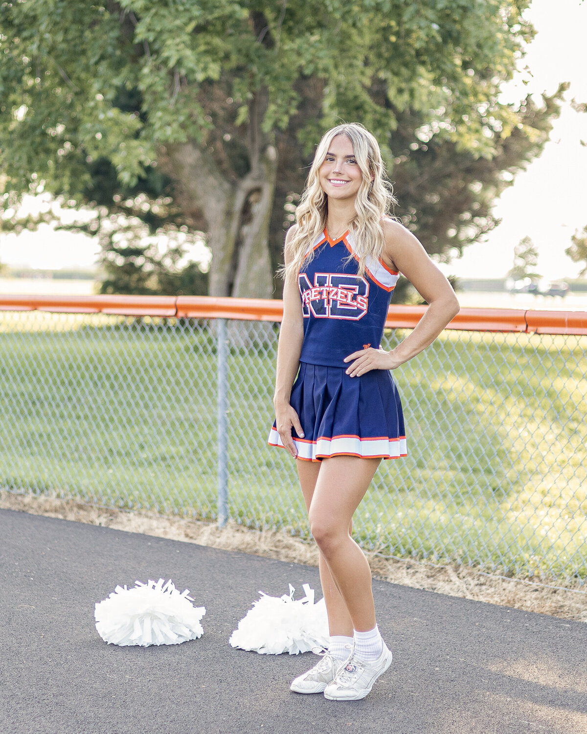 A high school senior female wearing a blue and orange cheerleading outfit, white shoes, smiling at the camera, standing on asphalt with poms next to her and hand on her hip, with long blonde hair