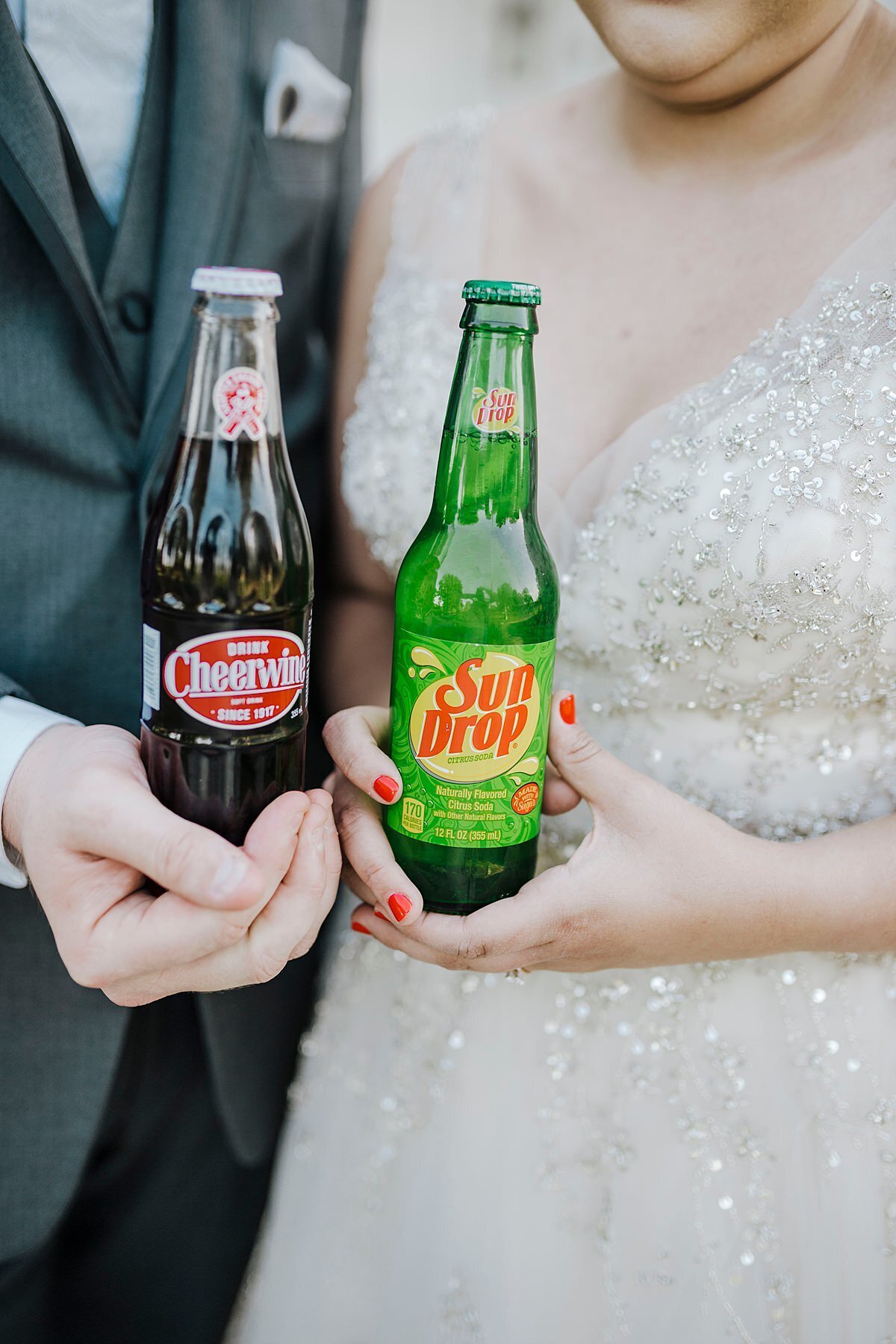 A bride wearing a sequined and beaded wedding dress with a plunging neckline and red nail polish holds a green glass bottle of Sun Drop soda. The groom, standing next to the bride in a dark gray suit holds a glass bottle of Cheerwine soda.