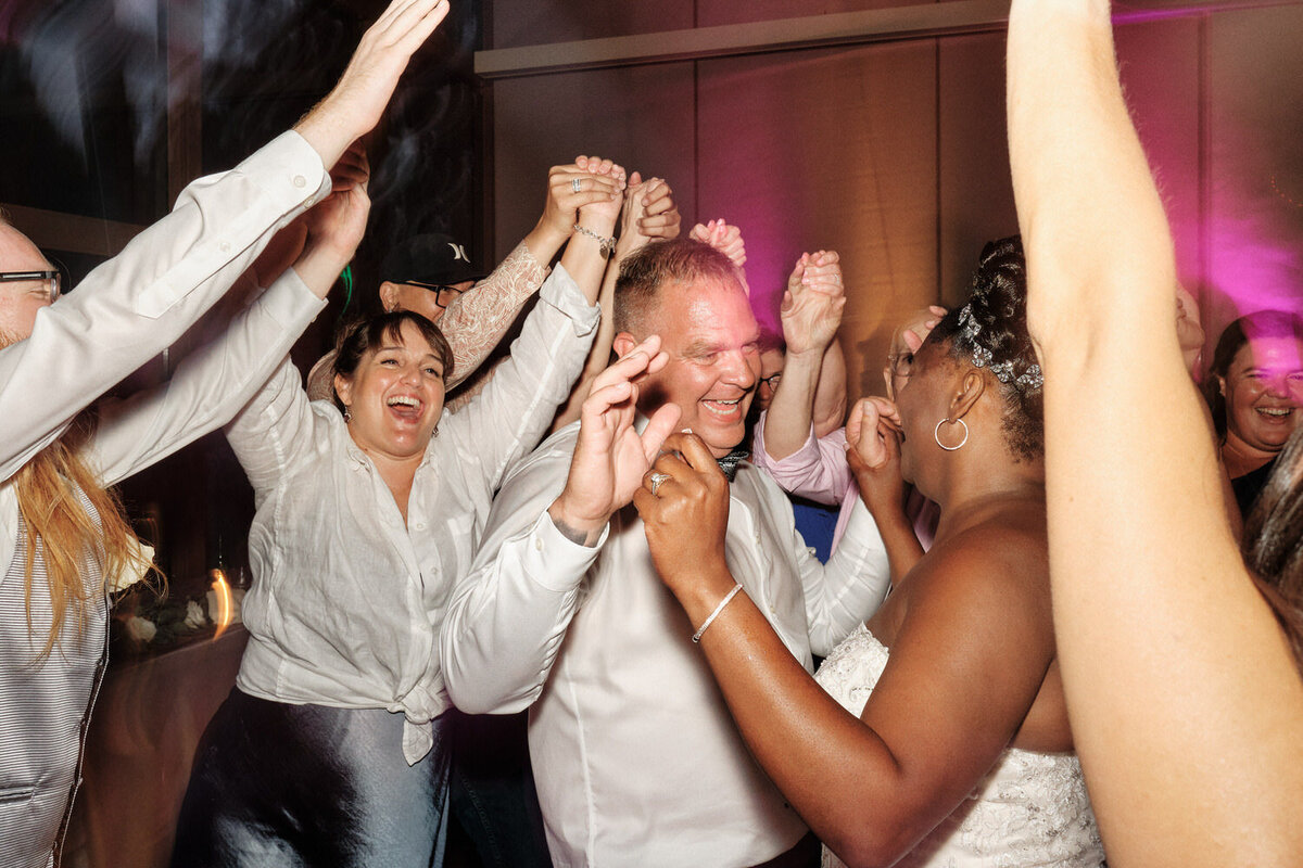 Groom laughs on the crowded dance floor while wedding guests throw their hands up