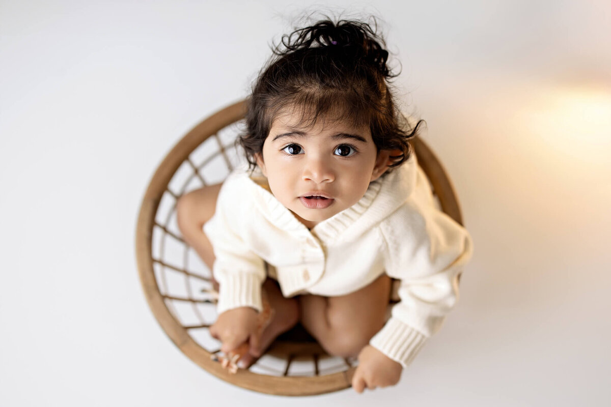 A young child with dark hair and big eyes sits in a round, woven basket, looking up at the camera. Captured by a Bloomington Illinois cake smash photographer, the child wears a cozy white sweater and is barefoot against a clean, bright white background.
