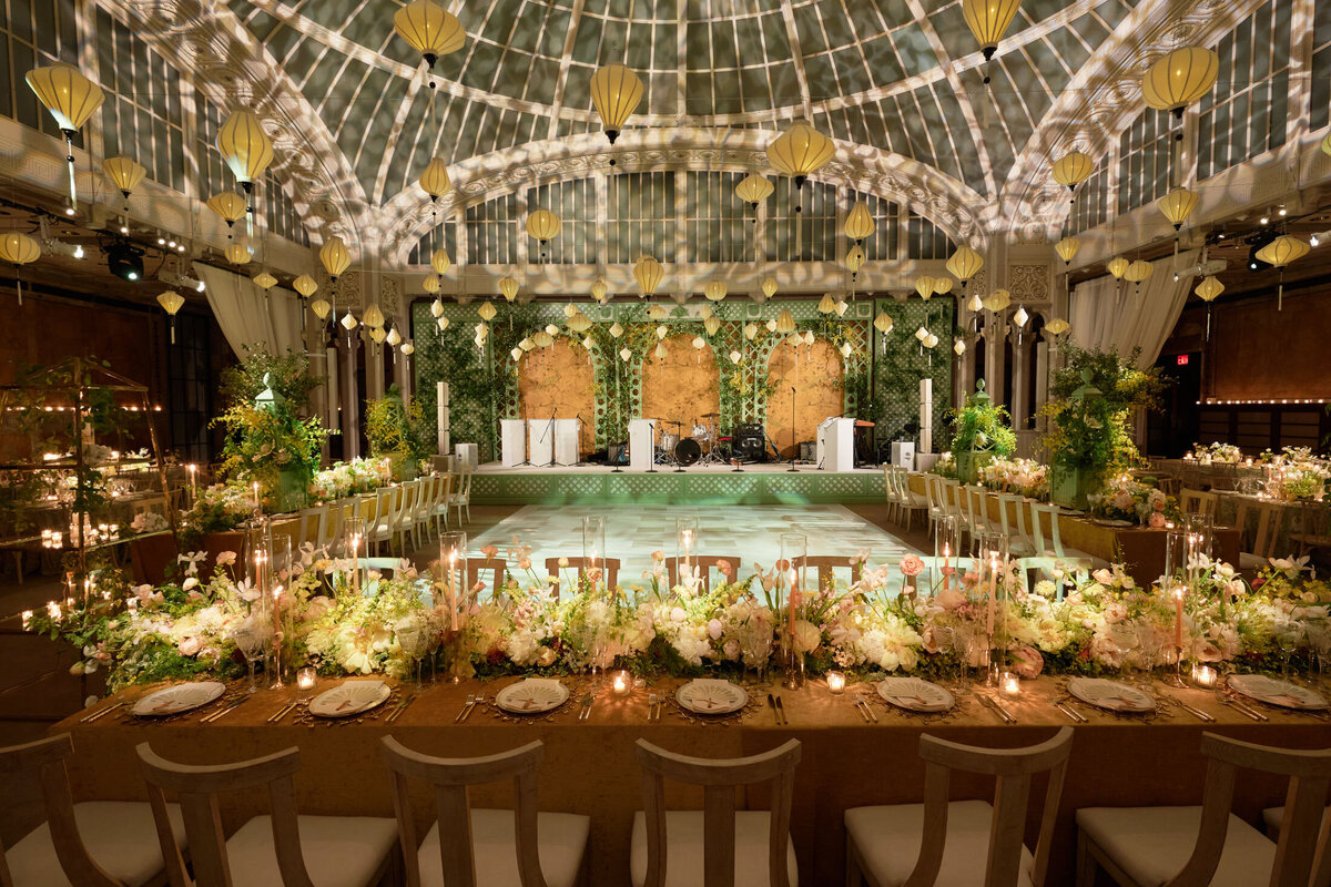 The photo was taken at the New York City, The New York Public Library by photographer Shawn Connell. It captures a lavishly decorated wedding reception with long tables adorned with elegant floral arrangements and candles. The spacious venue features a domed ceiling, hanging lanterns, and a dance floor, creating a luxurious and romantic ambiance. Videographers from Aaron Navak Films captured the beautiful moments of the event.