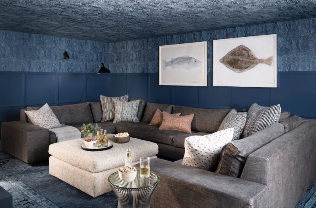 Boston interior design creates a navy blue media room with wainscot, wallpaper, a grey sectional, Walter G textile pillows, and large art for a stylish space.