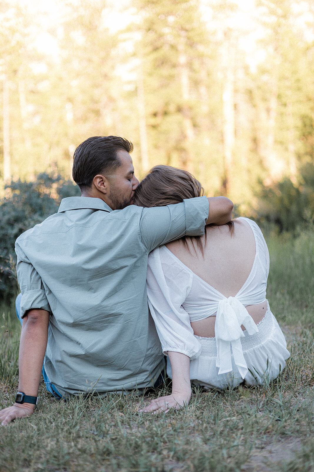 The man embraces the woman while they sit on the grass, her back to the camera; he places a gentle kiss on her head.